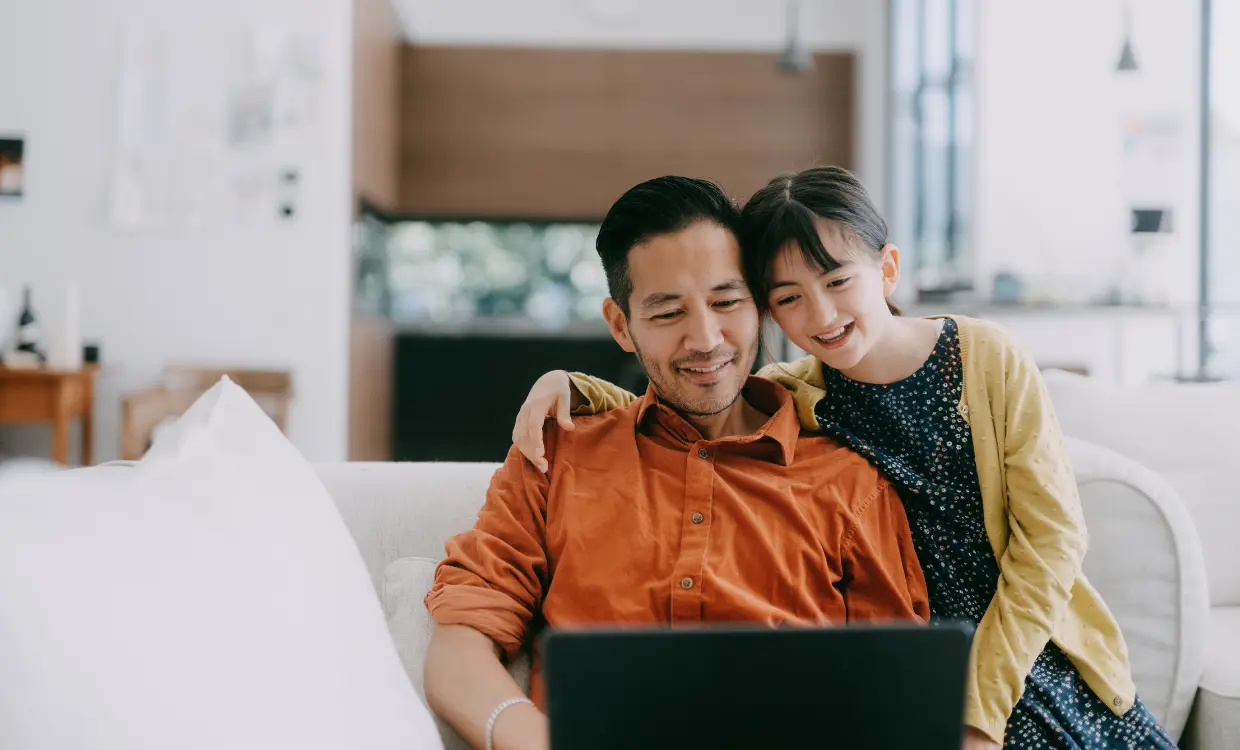 Father and daughter smile at a computer while sitting on the couch