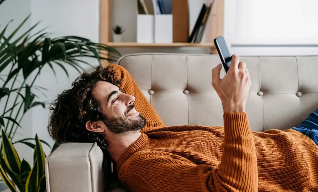 Man grins at his phone while lying on the couch