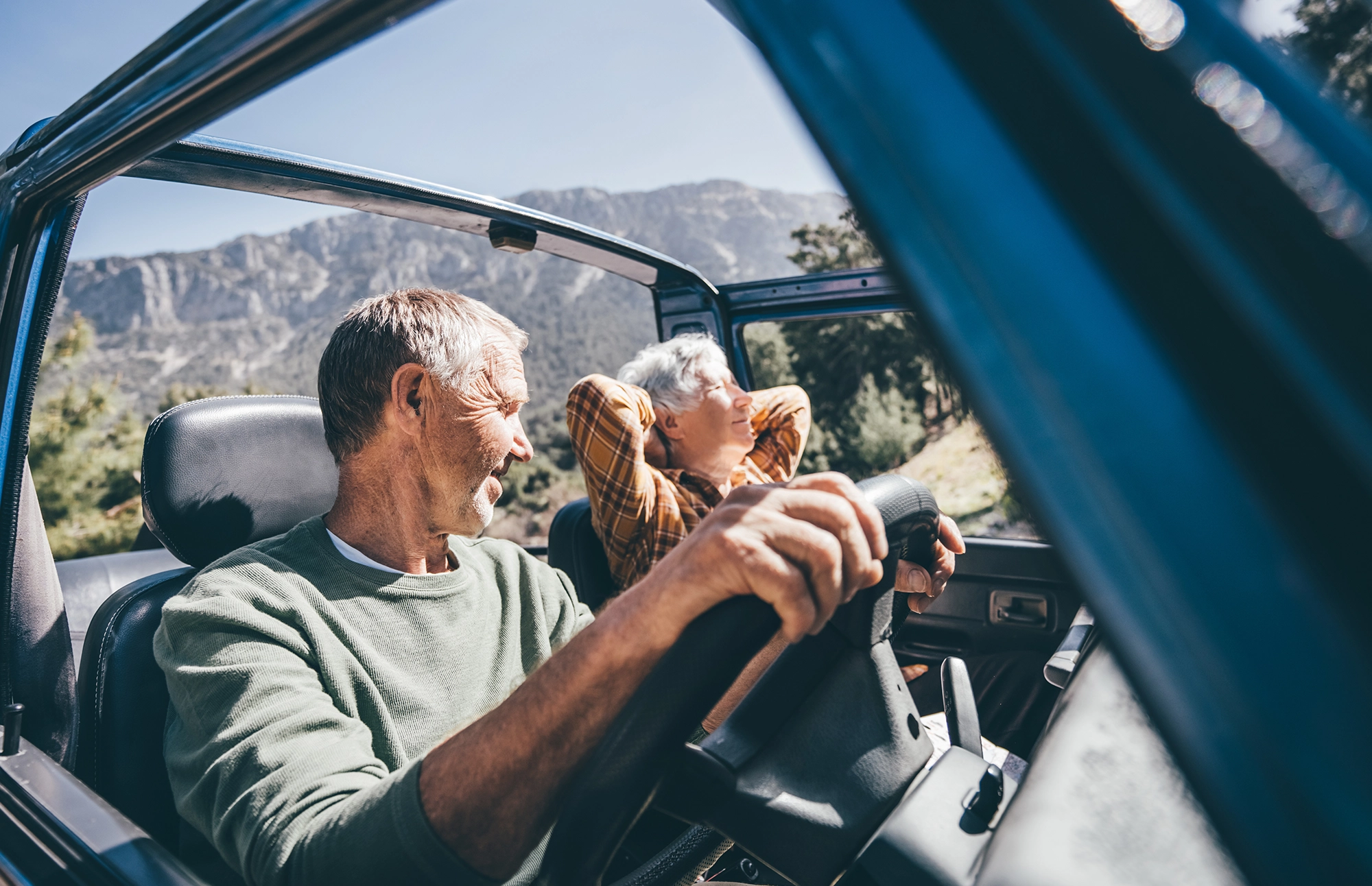 Older couple enjoying a scenic drive together in an open-top vehicle with mountains in the background.