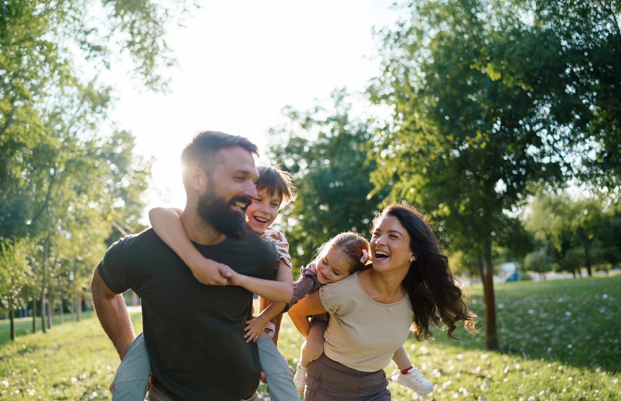 Family of four laughing together outdoors while giving their children piggyback rides in a sunny park.