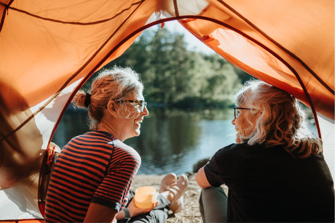Two older women smiling and chatting by a peaceful lake, sitting at the edge of an orange tent during a camping trip in nature.
