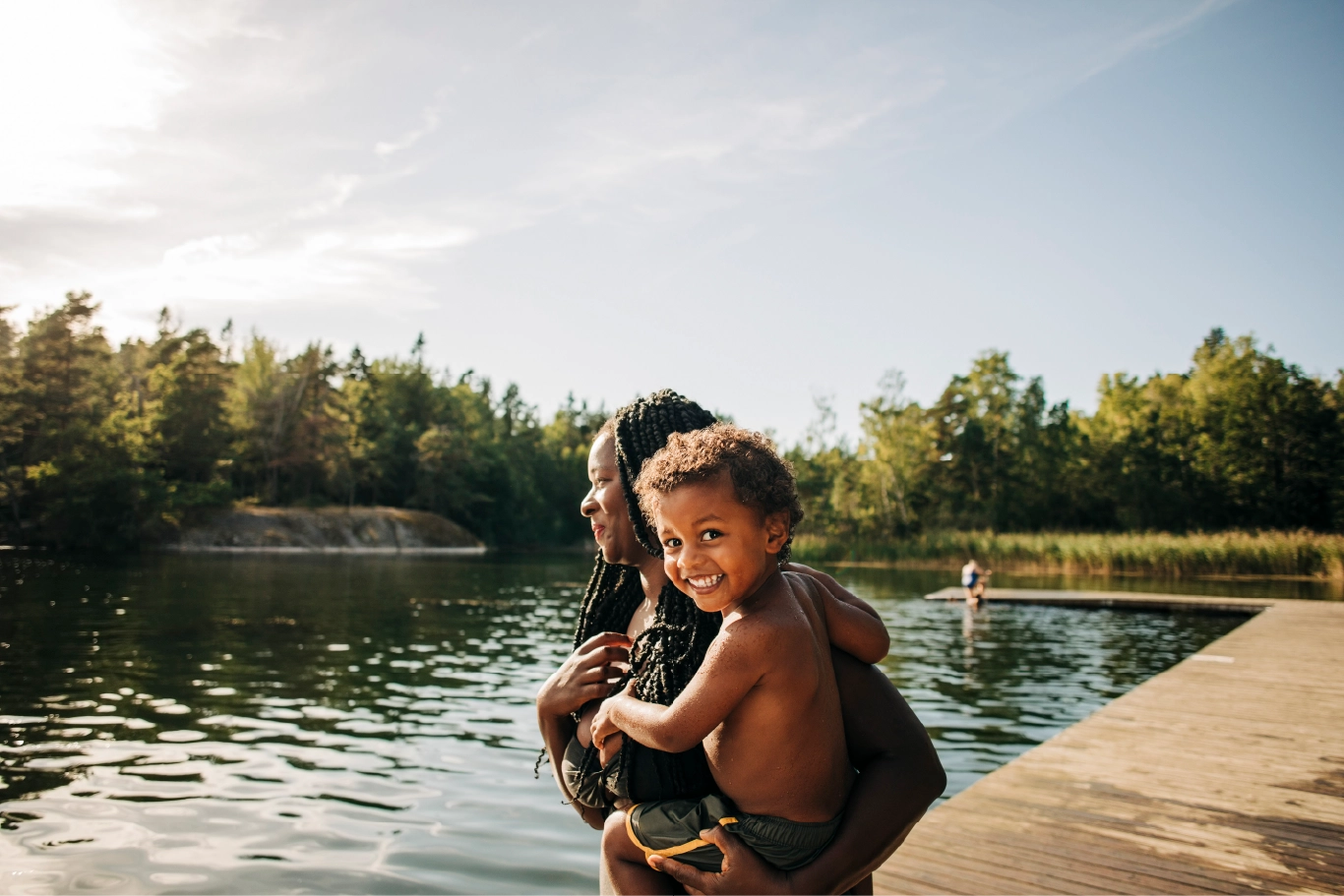 Smiling child held by parent at a lakeside dock on a sunny day, enjoying outdoor summer fun in nature.