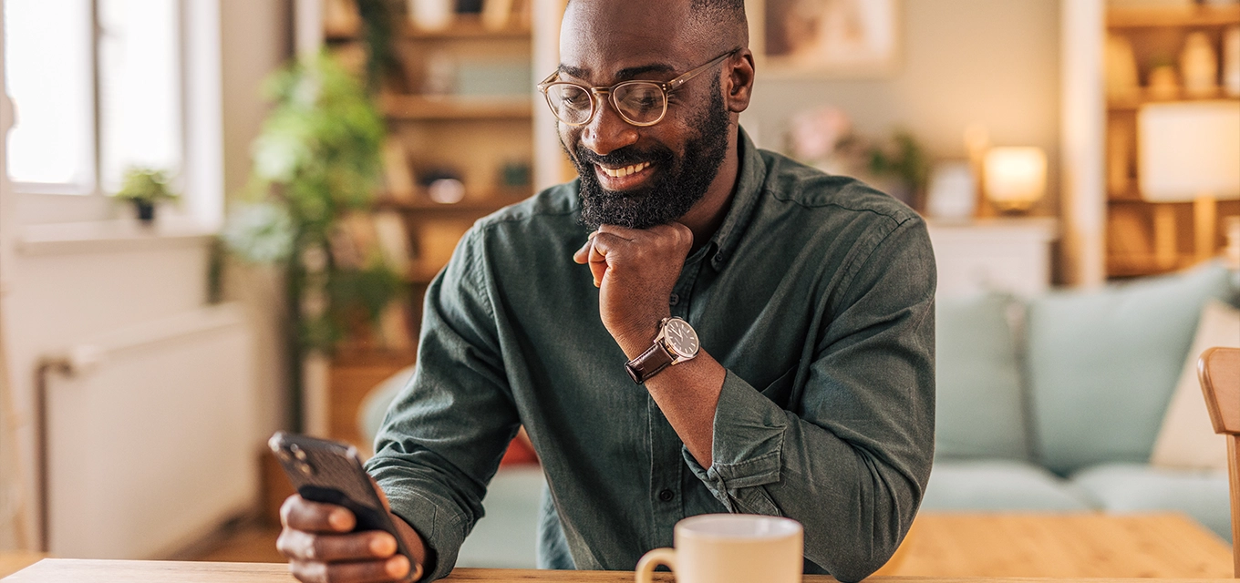Man smiling at his phone while sitting at a table with a coffee mug in a cozy home setting.