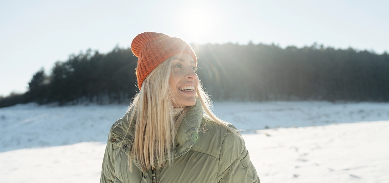 Person wearing a green winter jacket and orange knit hat smiling outdoors in a snowy landscape.