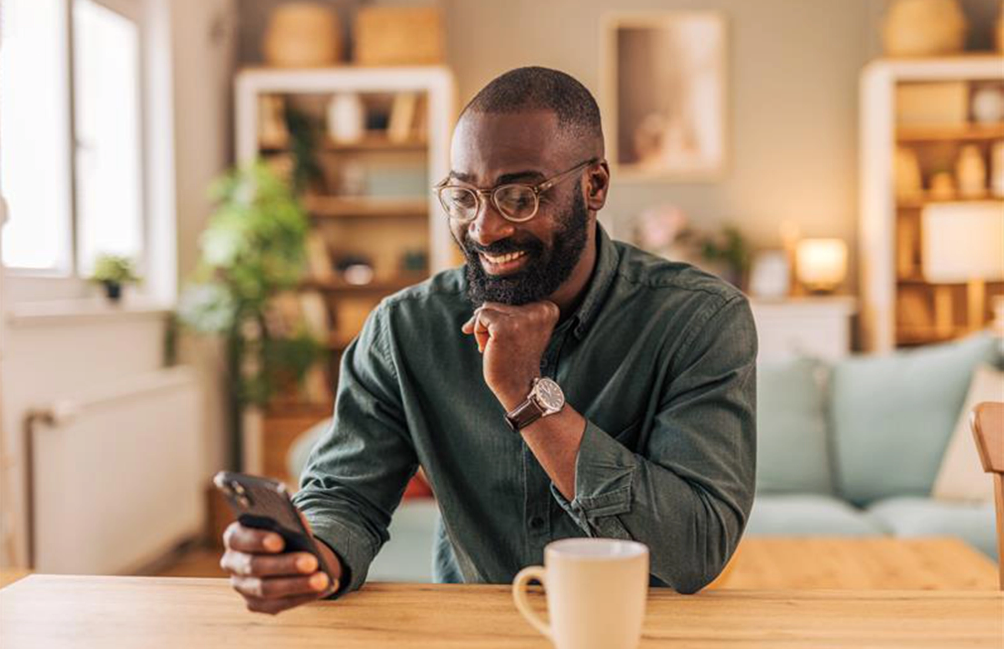 Man smiling at his phone while sitting at a table with a coffee mug in a cozy home setting.