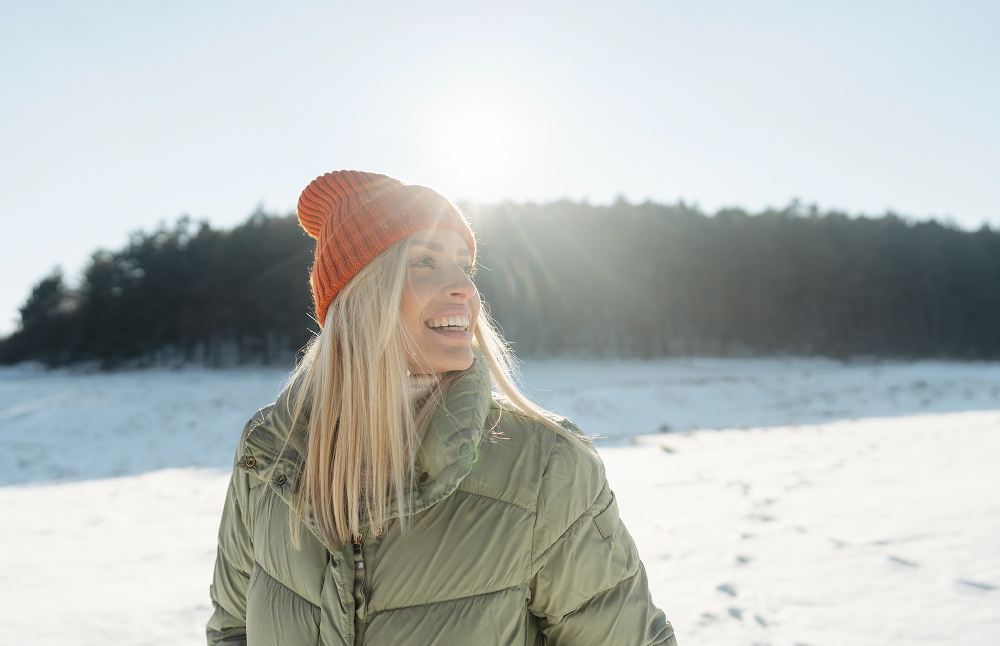 person wearing a green winter jacket and orange knit hat smiling outdoors in a snowy landscape.