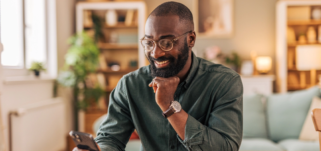 Man smiling at his phone while sitting at a table with a coffee mug in a cozy home setting.