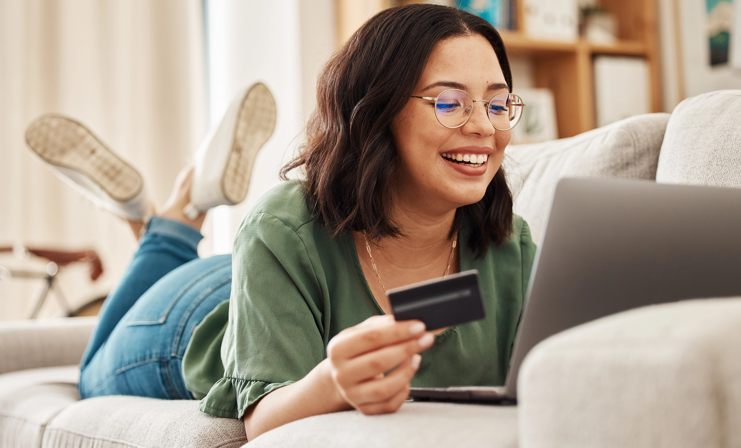 Woman smiles at computer while entering her card information to purchase an item while lying on the couch