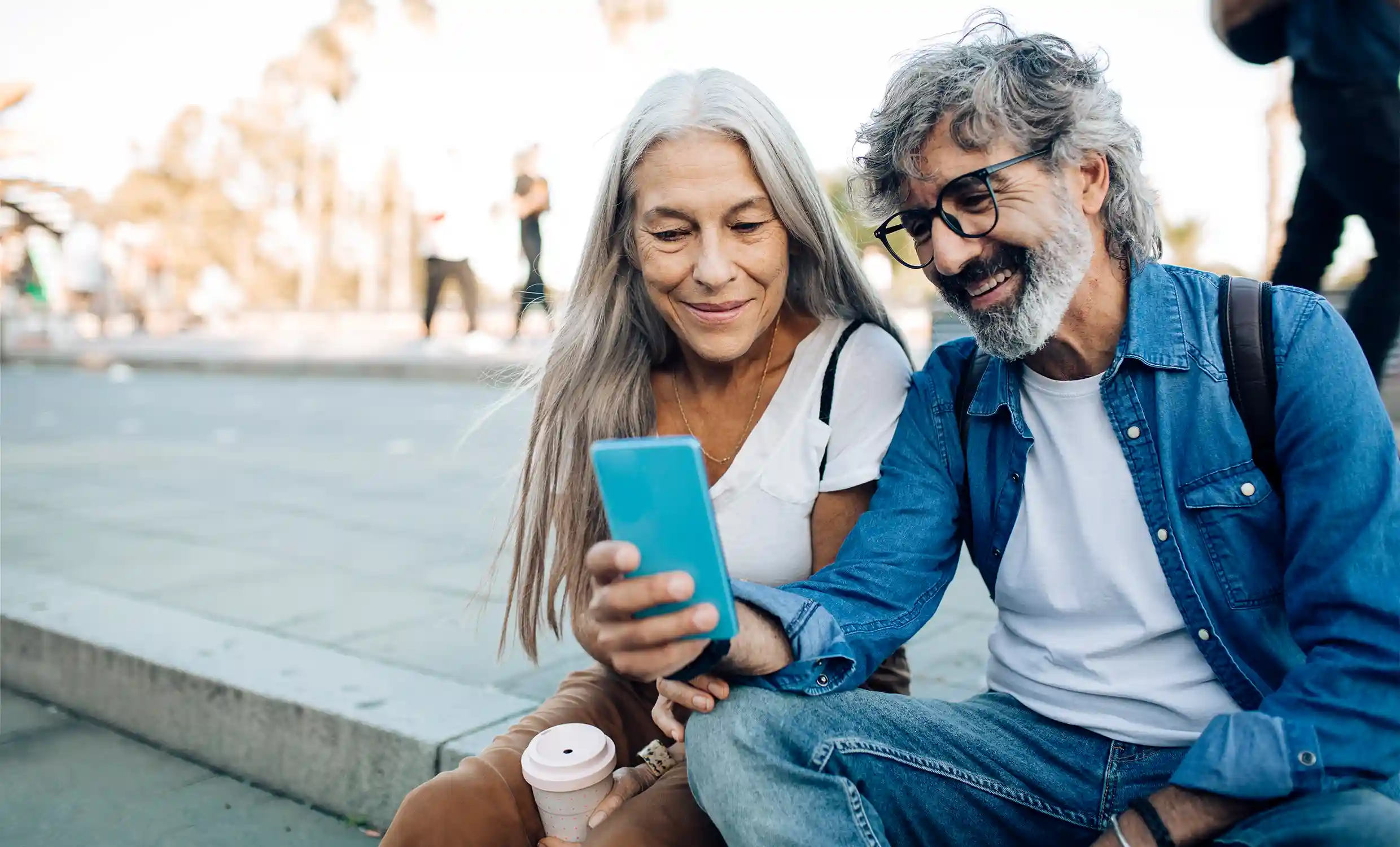 An older couple sit outside on a curb while smiling together at something they see on the man's cell phone