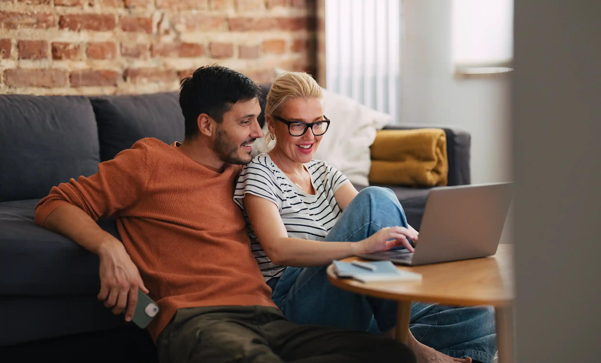 A man and woman sit together on their living room floor looking at a laptop on their coffee table