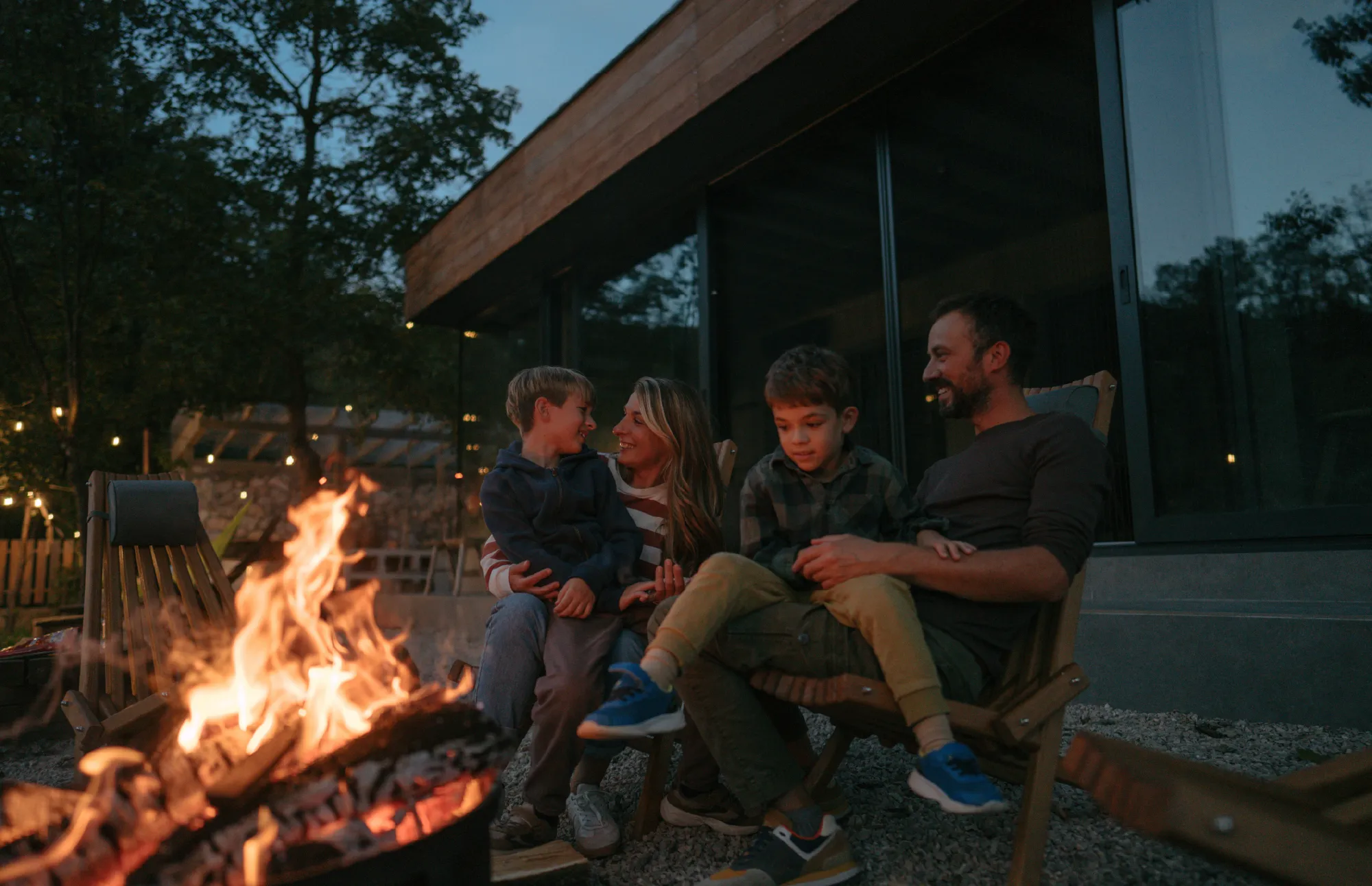 Family sitting together around a campfire outside a modern cabin at dusk.
