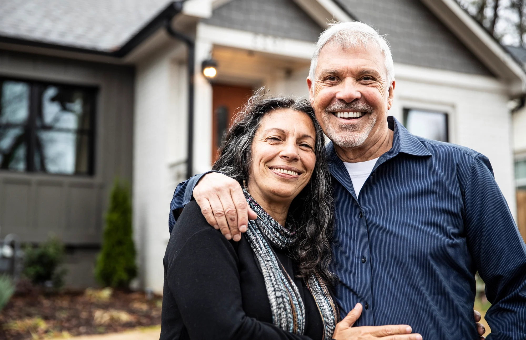 Man and woman smile at camera while hugging in front of their home