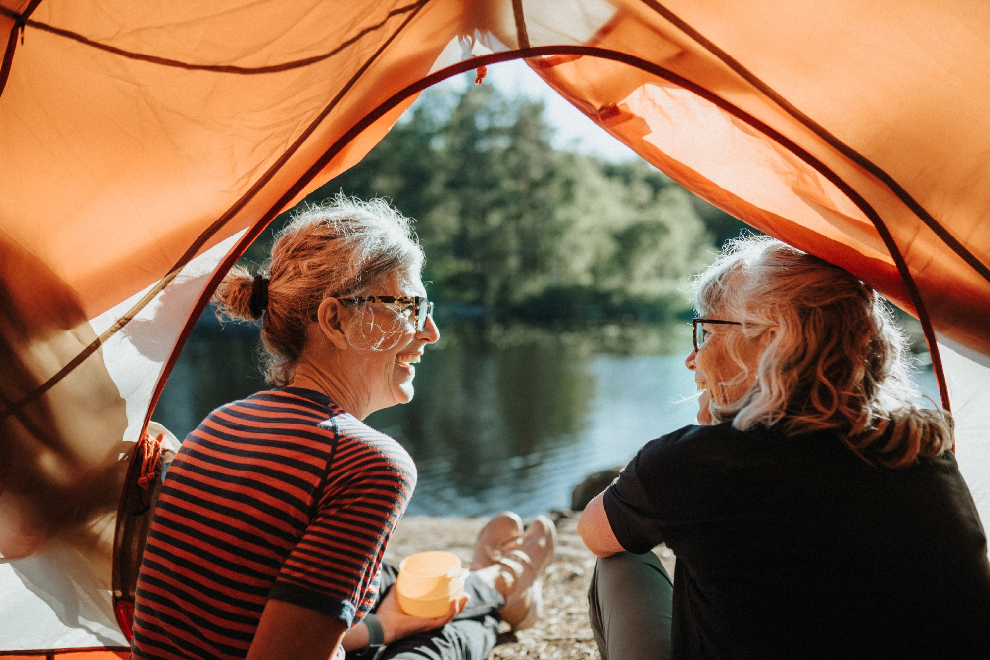 Two older women smiling and chatting by a peaceful lake, sitting at the edge of an orange tent during a camping trip in nature.