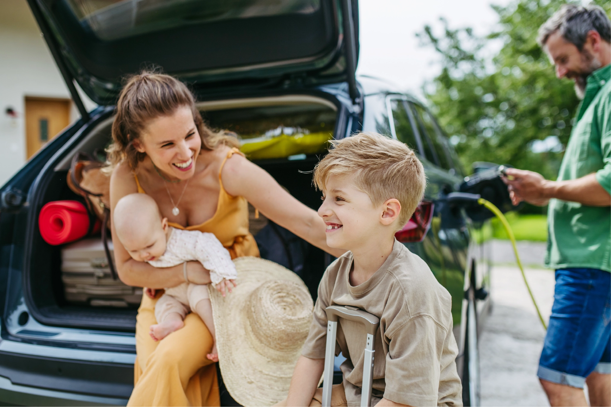Family getting ready for a road trip with packed luggage and electric car charging. Mom with baby, smiling boy with suitcase, and dad charging EV in driveway.