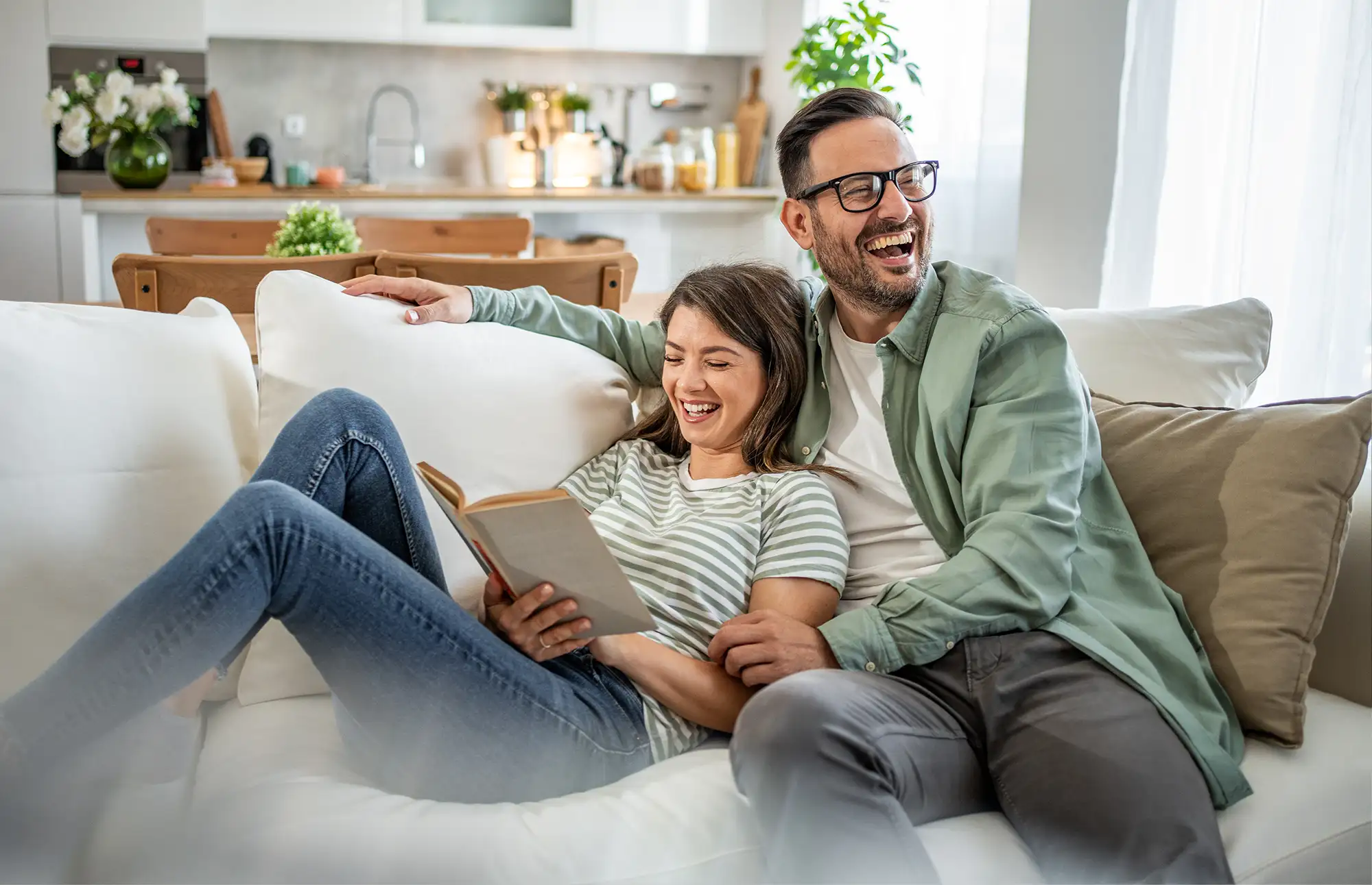 Couple lounge together on their couch and laugh at something the woman reads from her book