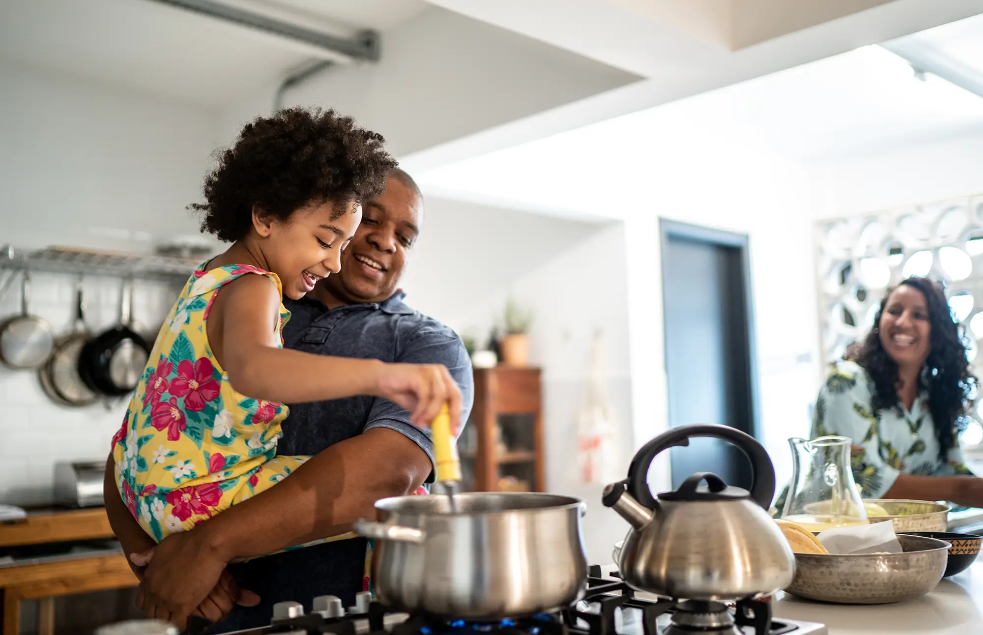 Family of three cooks dinner together in their kitchen with a young girl being held by her father stirring a pot