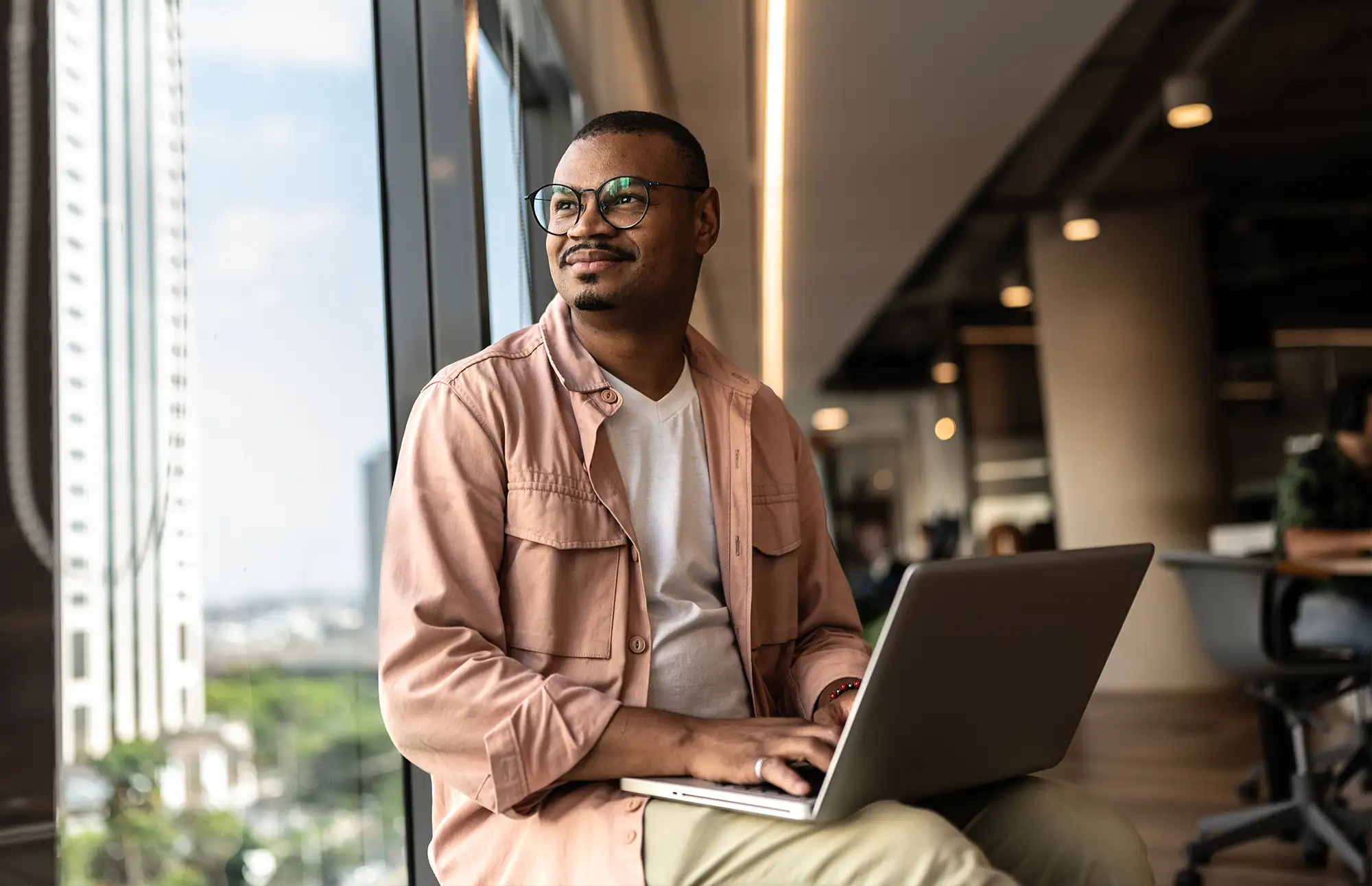 African American man sits by a window with a laptop in his lap while staring into the distance