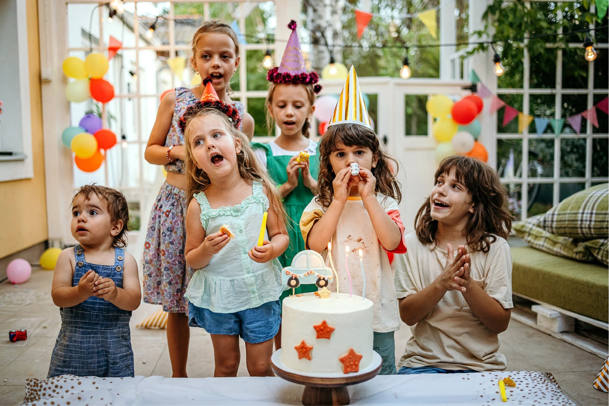 Group of young children celebrating a birthday with party hats, balloons, and a decorated cake, capturing joyful moments and family celebration at home.
