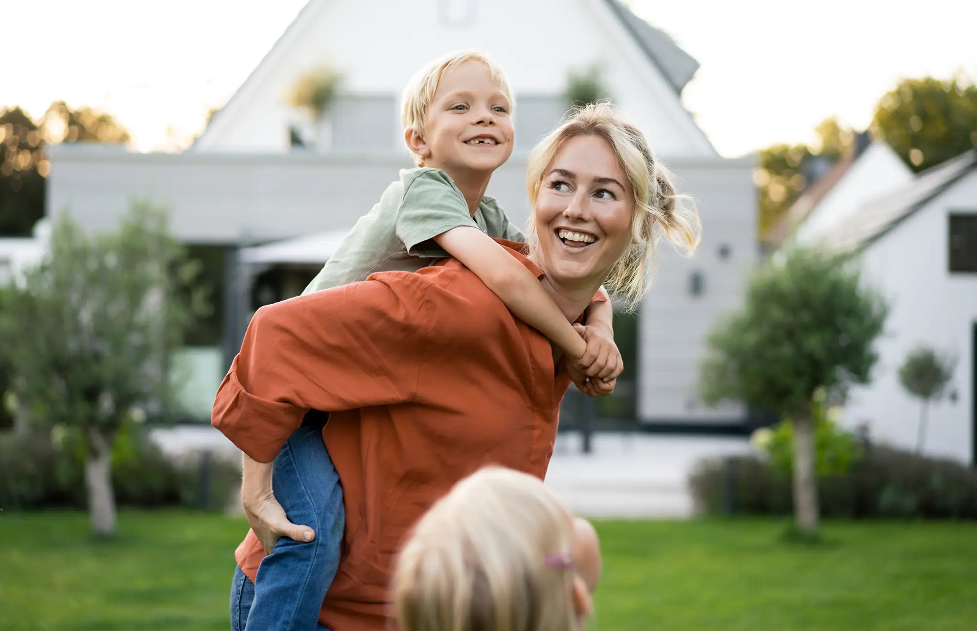 Blonde mother gives one of her children a piggyback ride in their yard while another child looks on