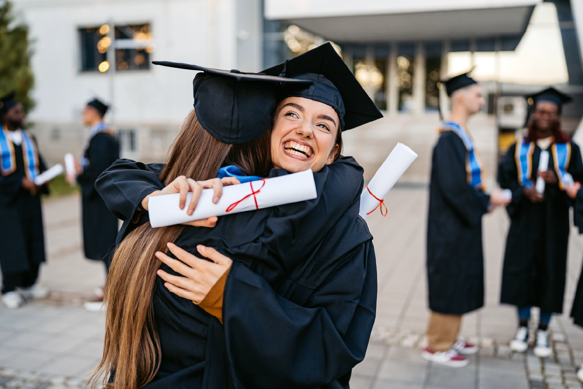 Two female students in graduation gowns embracing outdoors on a university campus, celebrating their achievements.