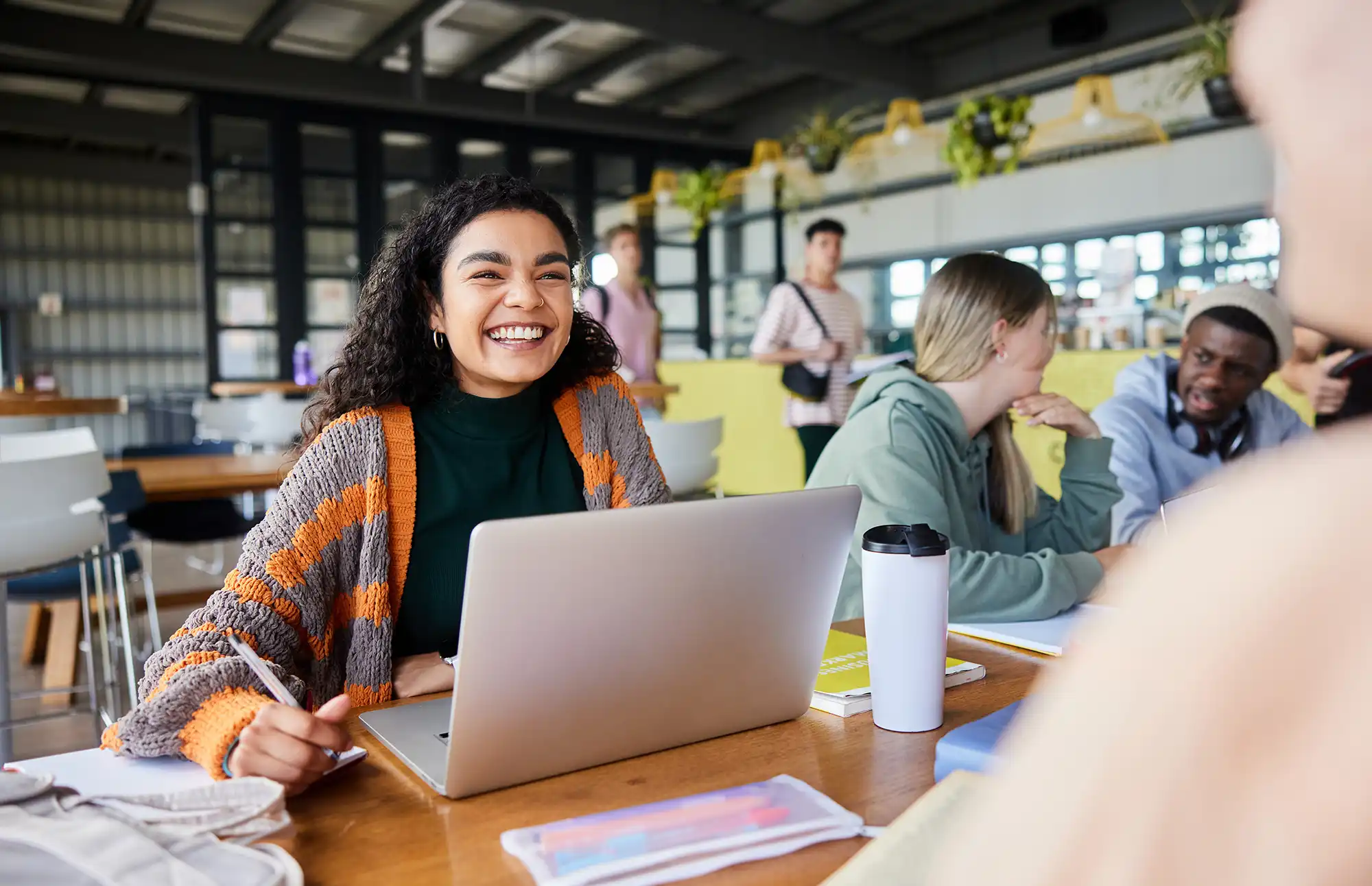 Young woman sits with other students in dining hall surrounded by books, coffee, and her laptop