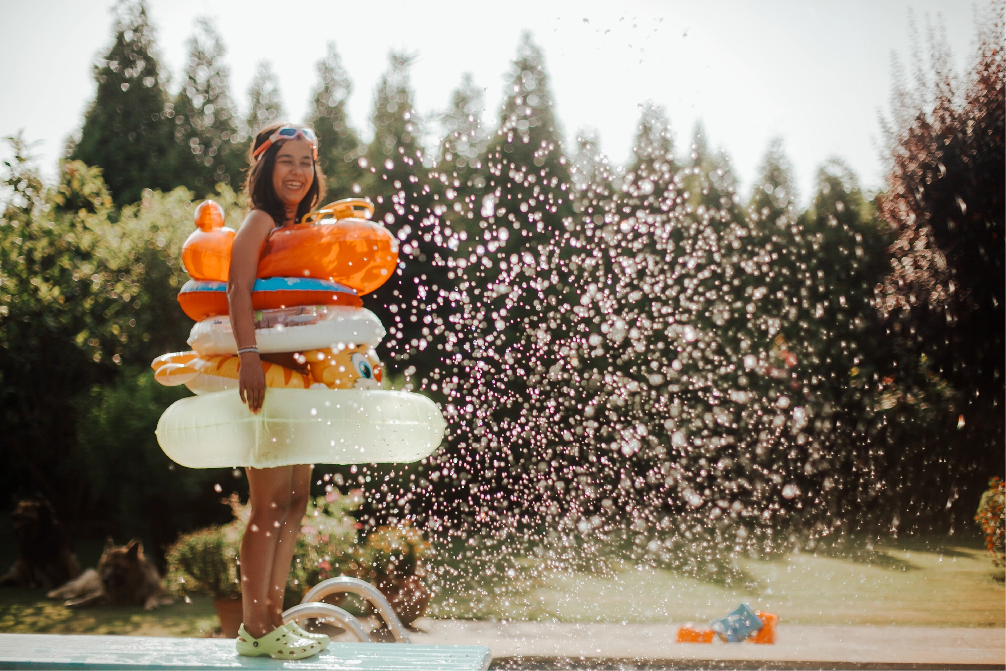 Smiling child wearing multiple colorful pool floats by a backyard pool with water splashing on a sunny summer day.