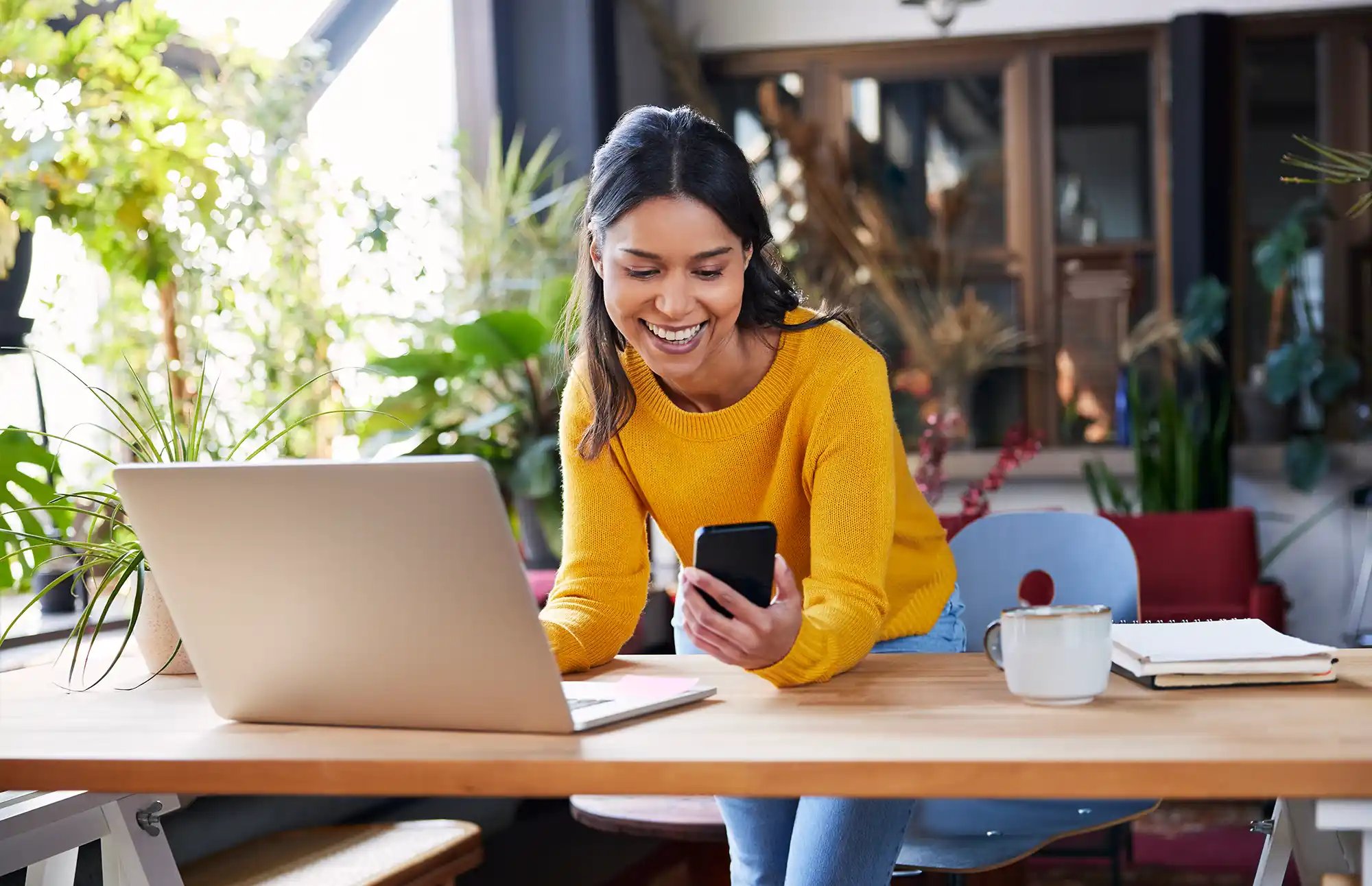 Woman stands over her desk in front of her laptop while turned to the side smiling at her cell phone