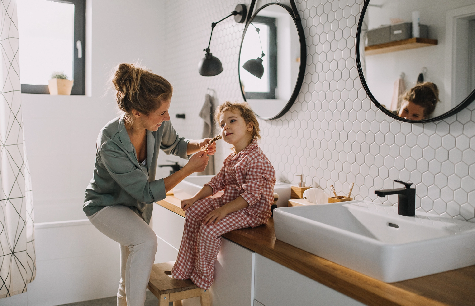 Adult helping a young child brush their teeth in a bright, modern bathroom.