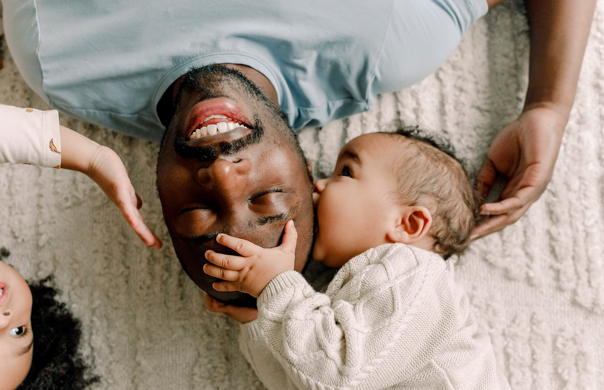 Adult smiling while lying on the floor with a baby reaching toward their face.