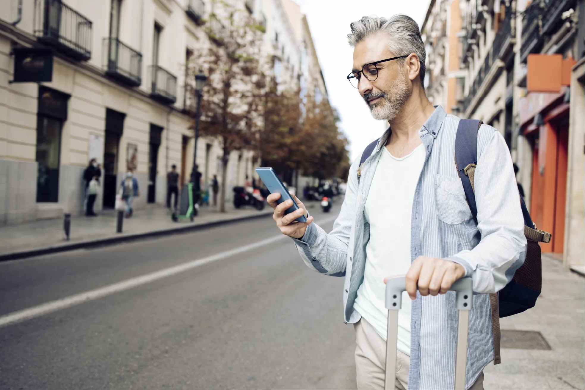 Man standing on a city street with a suitcase, looking at his phone.