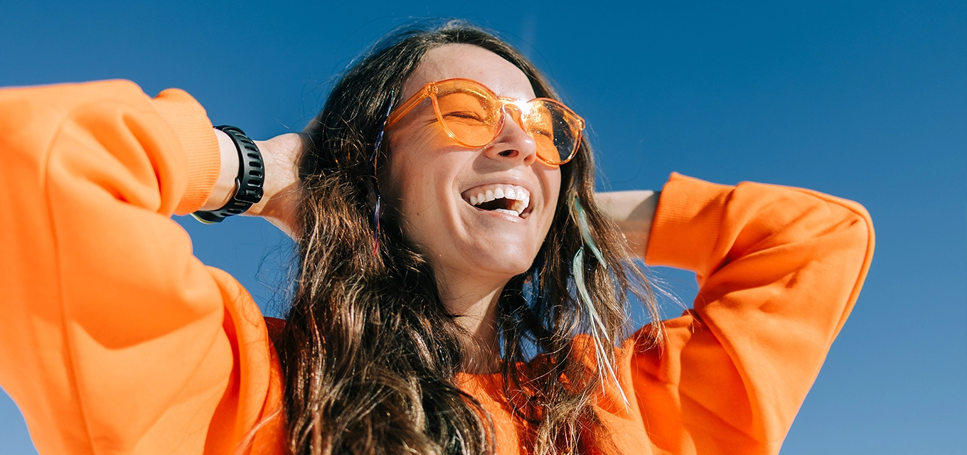 Woman smiling outdoors in orange sunglasses and a bright orange sweatshirt against a blue sky.
