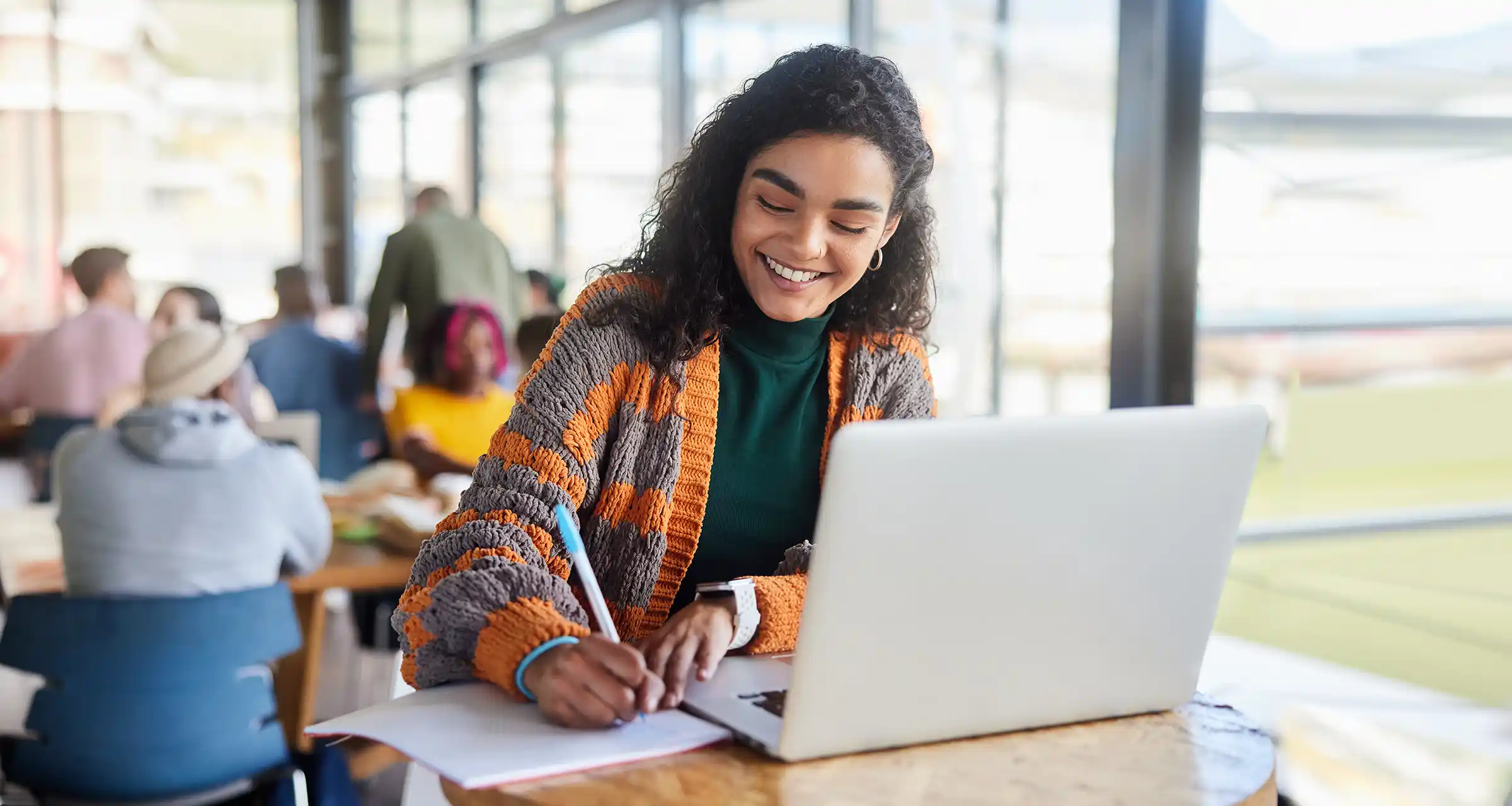 A young woman sits in a student lounge with a laptop and smiles while writing on a notepad