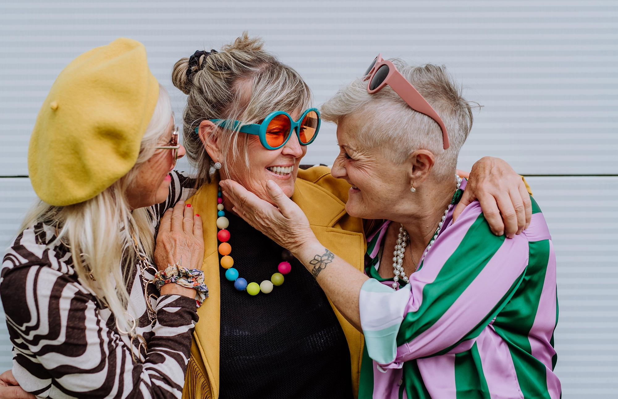 Three older women smiling and hugging while wearing colorful outfits and sunglasses.