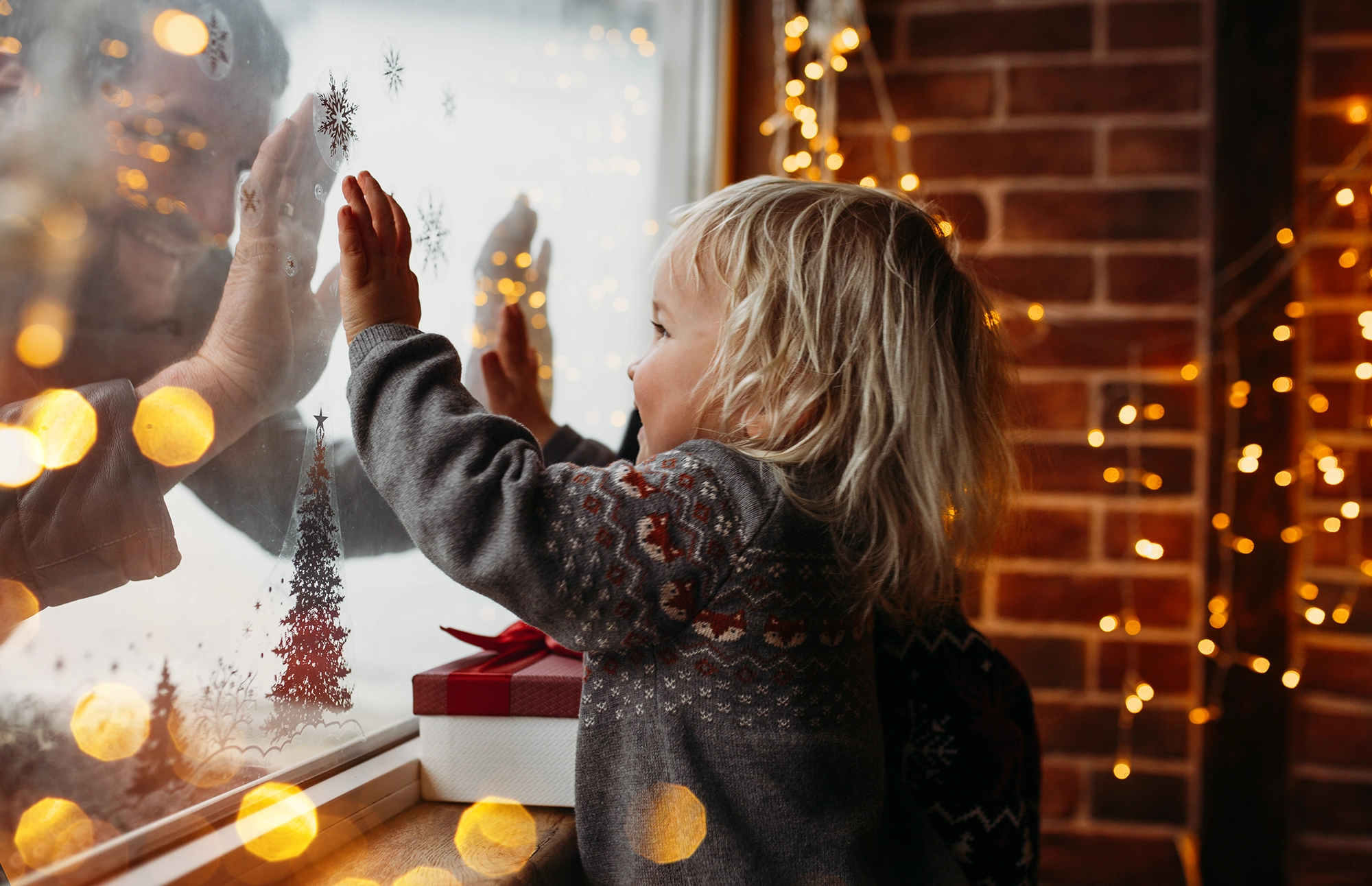 Child standing by a window touching the glass while holiday lights glow nearby.
